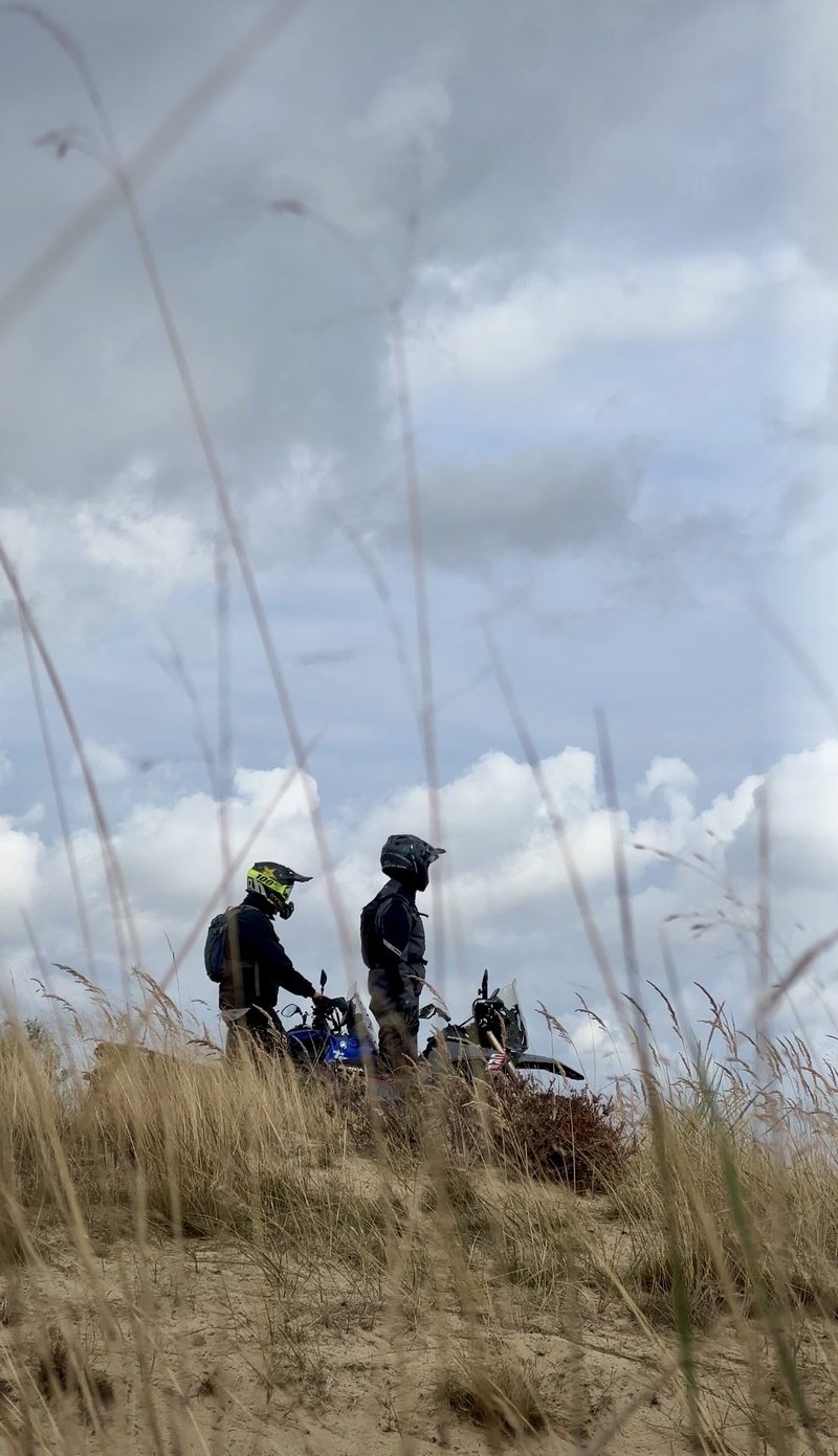 Kaks mootorratturit kiivritega liivasel küngal kõrge rohu vahel, taustal pilvine taevas